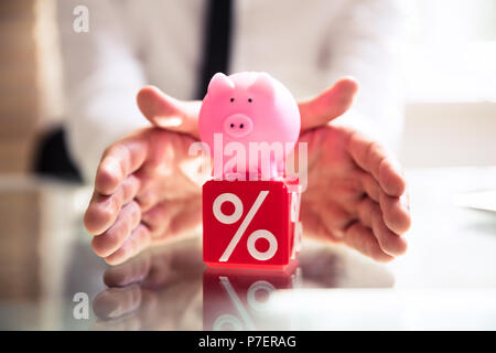 Close-up di mano di una persona di protezione Piggybank rosa e rosso Orologio cubico con simbolo di percentuale Foto Stock