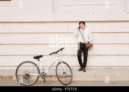 Giovane ragazzo bello con una bicicletta appoggiata contro la parete moderna in strada. Egli è in possesso di un telefono cellulare in mano. Foto Stock