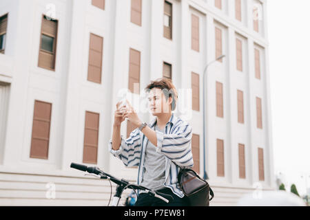 Elegante uomo chattare mentre si utilizza il telefono cellulare mentre è seduto sulla bicicletta, all'esterno. Vestito in plaid shirt, t-shirt e jeans. Close-up. Foto Stock