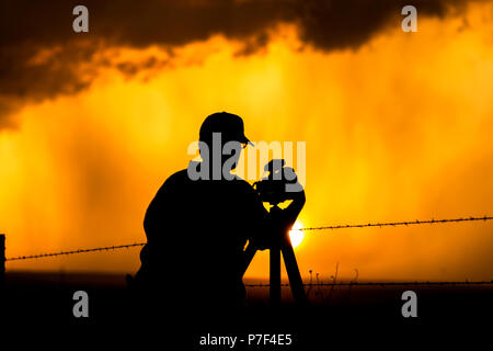 Un fotografo è incorniciato contro un temporale tramonto mentre la pioggia si riversa nel lontano arancione e tonalità gialla. Foto Stock