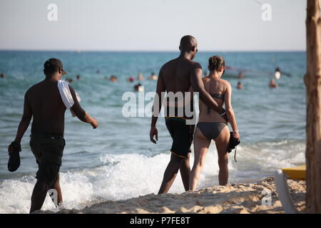 Grande uomo africano passeggiate abbracciando la ragazza europea sulla riva in Hammamet, Tunisia, essa abbracci lui, essi a piedi attraverso la schiuma delle onde all'inizio serata estiva Foto Stock