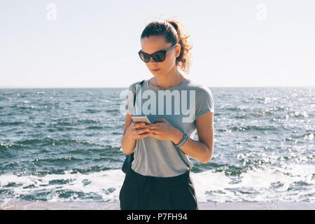 Moda giovane donna in occhiali da sole texting permanente sullo sfondo del mare e sole flare. Femmina blu che indossa una maglietta e pantaloni neri utilizzando smart Foto Stock