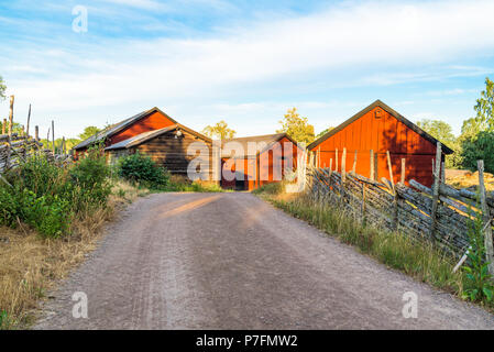 La ghiaia country road passando alcuni granai su una sera d'estate. Tradizionale scandinavo roundpole recinzioni sui lati della strada. Ubicazione Villa Stensjo Foto Stock