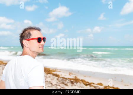 Primo piano della giovane bello attraente uomo, volto in tshirt, t-shirt sorridente sulla spiaggia durante la giornata di sole con occhiali da sole rosso in Florida ocean Foto Stock