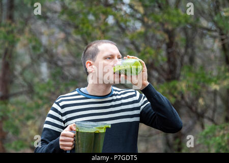 Giovane uomo in piedi fuori, all'aperto, tenendo in plastica contenitore blender, bere dal bicchiere per frullato verde realizzato da vegetali verdi Foto Stock
