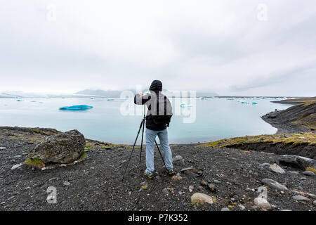 Retro del giovane fotografo in inverno freddo rivestire fotografare fotografare ghiacciaio blu iceberg nel lago laguna in Islanda su treppiede Foto Stock