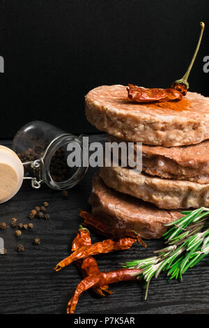 Materie tritata burger di carne con erbe e spezie preparato per la cottura alla griglia con rosmarino, caldo rosso peperoncino, pepe nero su nero tavolo in legno Foto Stock