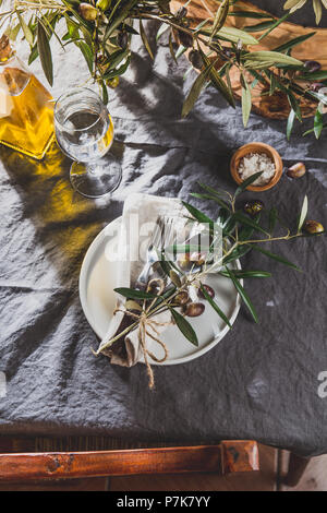 Impostazione tabella con lino grigio Tovaglia e tovagliolo, piastra bianca, posate e ulivo decorazione di diramazione e olio di oliva. Vista dall'alto. Foto Stock