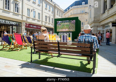 Bath, Regno Unito, 7 luglio, 2018. Come bagno gode di un altro molto calda e soleggiata giornata gli acquirenti sono ritratte godendo il sole in SouthGate shopping center dove un grande schermo è stato impostato in modo che gli appassionati di tennis possono guardare il torneo di Wimbledon Tennis Championship, forecasters prevedono che il clima caldo proseguirà anche nella prossima settimana. Credito: lynchpics/Alamy Live News Foto Stock