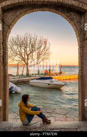 Tourist ammirando la luce del tramonto sul porticciolo di Punta San Vigilio dall' arco dell'incantevole risalente al XVI secolo inn sulla sponda orientale del Lago di Garda, provincia di Verona, regione Veneto, Italia. Foto Stock