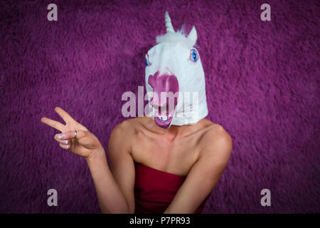 Funny Girl unicorn flirta e gesticulating. Freaky giovane donna in maschera comica sorge su sfondo viola. Ritratto di signora inusuale in abito rosso. Foto Stock