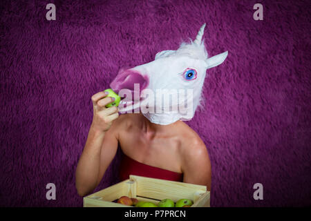 Freaky giovane donna in maschera comica sorge sullo sfondo viola e mostra la cassa di frutta. Funny Girl unicorn eat apple. Ritratto di signora inusuale. Foto Stock