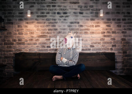 Insolito giovane uomo nella maschera di comico si siede da solo sul letto in un elegante appartamento. Grave unicorn in abiti casual. Freaky uomo a casa. Foto Stock