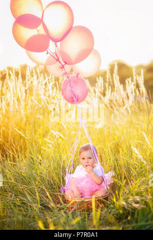 Bellissima bambina in abito rosa seduta sul cestello con palloncini all'esterno. Celebrazione del primo concetto di compleanno Foto Stock