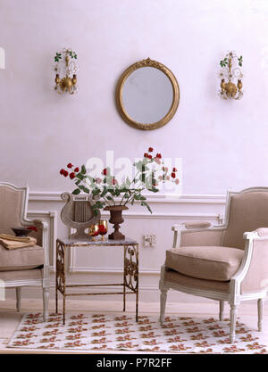 Grigio verniciato imbottiti in stile francese di sedie e piccolo tavolo in metallo in un economia di salotto in stile con le luci a parete su entrambi i lati dello specchio Foto Stock