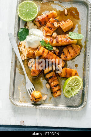 Vassoio di caramellato ananas grigliato con gelato, menta e i dadi sulla luce di una superficie di legno, vista dall'alto Foto Stock