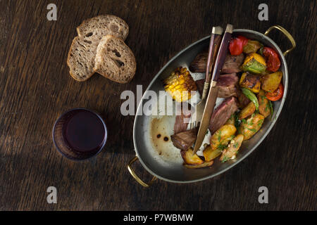 Bistecca con patate fritte e verdure in padella su sfondo scuro con un bicchiere di vino rosso, vista dall'alto. Tavola di Cena concept Foto Stock