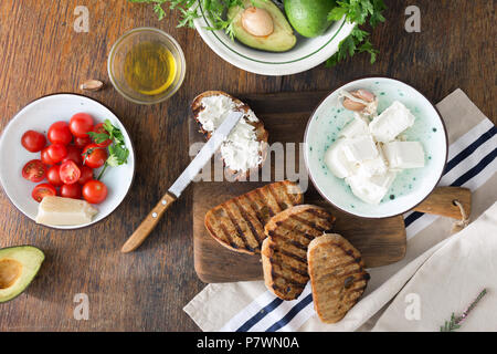 Gli ingredienti per cucinare bruschetta con formaggio feta, pomodorini e avocado su un tavolo di legno, vista dall'alto. Il cibo italiano Foto Stock