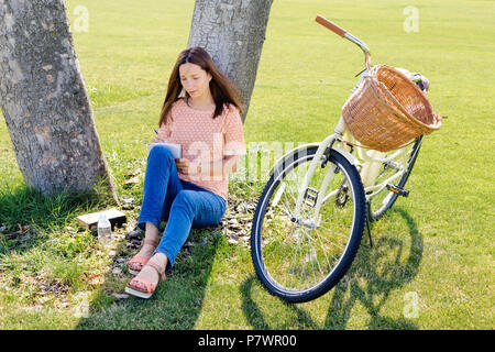 Immagine di donna giovane studente scrive in un notebook seduto sotto un albero in un giorno di estate Foto Stock