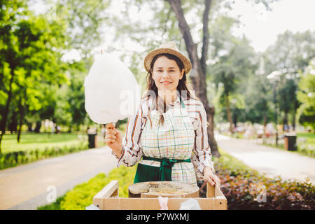 Foto tema small business la cottura di dolci. Una giovane donna caucasica con un grembiule commerciante nel cappello il proprietario dell'uscita fa un candy floss, una fiera Foto Stock