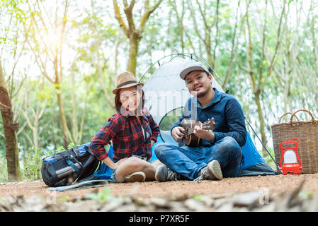 Asian giovane camping nella foresta. L'uomo gioca ukulele con la donna nella parte anteriore della tenda da campeggio. Lampada e cesto zaino e l'elemento. Persone e outdoor Foto Stock