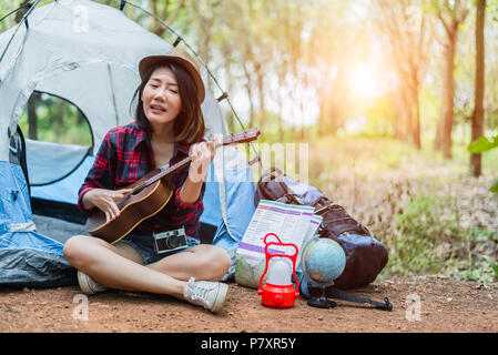 Bella donna asiatica giocando Ukulele nella parte anteriore della tenda da campeggio nei boschi di pino. Persone e stili di vita del concetto. Avventura e tema Viaggi. Foto Stock