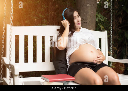 Asian giovane donna incinta rilassante e ascoltare musica su altalena nel parco giardino con i libri. Persone e concetto di intrattenimento. La felicità e il piacere c Foto Stock