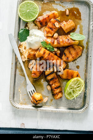 Vassoio di caramellato ananas grigliato con gelato, menta e i dadi sulla luce di una superficie di legno, vista dall'alto Foto Stock