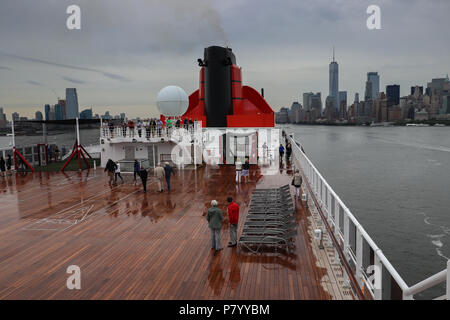 Passeggeri guarda la Queen Mary 2 lascia il porto di New York da un umido del piano superiore su una serata piovosa, sullo skyline di New York visibile in distanza. Foto Stock