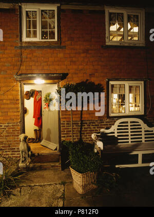 Bay Tree in pentola accanto al banco di Lutyens al di fuori del cottage di notte con finestre illuminate Foto Stock