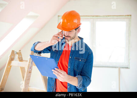 Ritratto di giovane tuttofare holding appunti nella sua mano ed effettuare una chiamata mentre si sta in piedi in zona di costruzione. Foto Stock