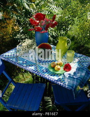 Close-up di calce-verde caraffa di vetro e bicchieri fatti a mano in blu+bianco piastrelle a mosaico e rabboccato Tavolo da giardino con sedia blu Foto Stock