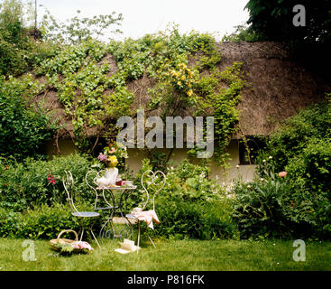 Tavolo in metallo e sedie sul prato di fronte fatiscenti cottage con piante rampicanti crescente sul tetto di paglia Foto Stock