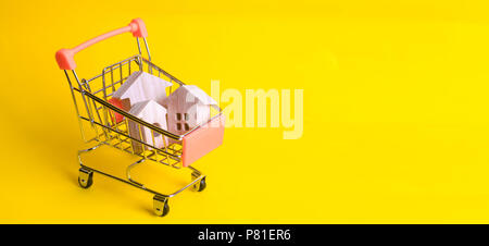 Un carrello per supermercati con case in legno si erge su uno sfondo giallo. Il concetto di home shopping e marketing, commercio e. Housin conveniente Foto Stock