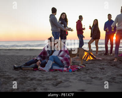 Coppia giovane seduto con gli amici intorno al falò sulla spiaggia al tramonto bevendo birra Foto Stock