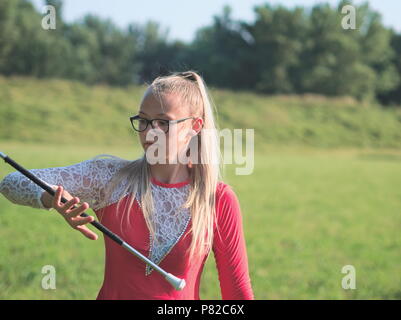 Bespectacled Blonde Teen Majorette ragazza fa roteare Baton all aperto in abito rosso Foto Stock