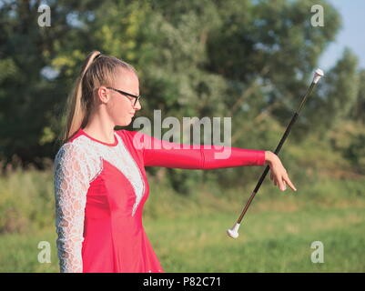 Bespectacled Blonde Teen Majorette ragazza fa roteare Baton all aperto in abito rosso Foto Stock