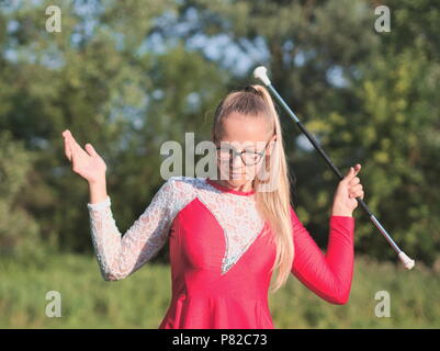 Bespectacled Blonde Teen Majorette ragazza fa roteare Baton all aperto in abito rosso Foto Stock