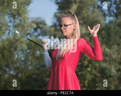 Bespectacled Blonde Teen Majorette ragazza fa roteare Baton all aperto in abito rosso Foto Stock