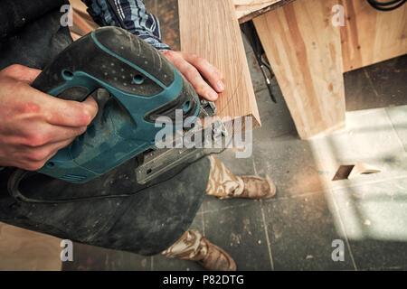 In prossimità di un falegname esperto in abiti da lavoro e piccole buiness proprietario carving è una tavola di legno su un moderno trapano a mano in un workshop di luce Foto Stock