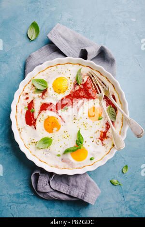 Uova cotte shakshouka con salsa di pomodoro, erbe aromatiche e basilico fresco in forma cotta sul tavolo blu. Pronti per la prima colazione. Vista da sopra Foto Stock