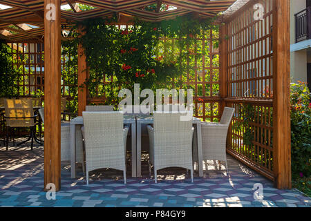 Un gazebo in legno con tavolo e sedie in vimini e piante con fiori di colore rosso Foto Stock