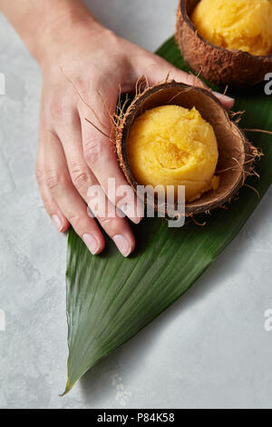 Un gelato al mango sorbetto in un guscio di noce di cocco è tenuto da una femmina di mano su una foglia di palma su un cemento grigio Sfondo. Vista superiore Foto Stock