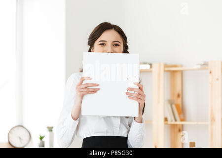 Giovane donna con sorpresa o felice occhi che spuntavano da dietro il poster di carta. Imprenditrice azienda big banner bianco in un ufficio moderno. Foto Stock