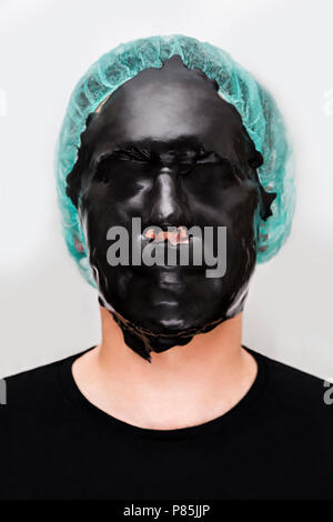 Paziente di sesso maschile con alginato di pelle alghe maschera nera sulla faccia su sfondo bianco. Foto Stock