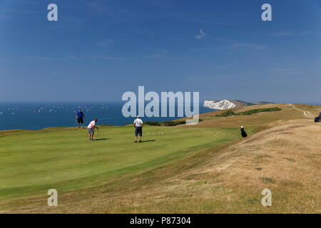 Gli amanti del golf putting su un verde a Freshwater Bay Golf Club con le coloratissime vele del giro dell'isola in barca in gara sullo sfondo l'Isola di Wight Foto Stock