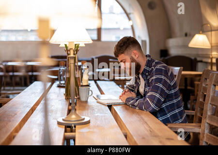 Giovane imprenditore barbuto siede in cafe, home a tavola e scrive in notebook, vicino si trova computer tablet. L uomo è lavorare, studiare. Foto Stock