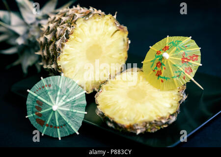 Fette di ananas maturo su una piastra nera con decorazioni per cocktails, ombrello su sfondo scuro Foto Stock