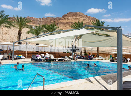 I turisti godono di una piscina all'aperto parzialmente ombreggiata in un hotel resort a Ein Bokek sul Mar Morto, con montagne desertiche e cielo blu Foto Stock