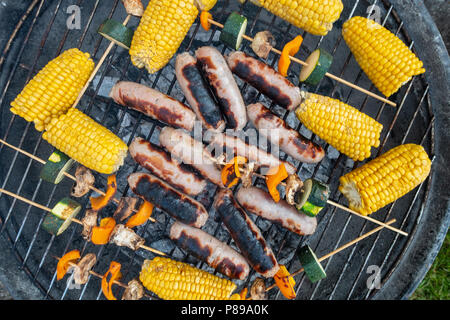 Salsicce e salami, sulla pannocchia di mais e spiedini di vegetali di funghi, il pepe e la zucchina cucinando fuori su un barbeque. Foto Stock